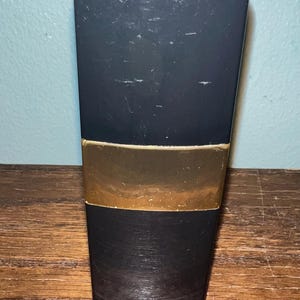 May include: A tall, rectangular black bottle with a smooth, matte finish and a prominent horizontal gold band across the center. The top of the bottle features a circular silver cap with an embossed logo design, and the base has a textured surface.