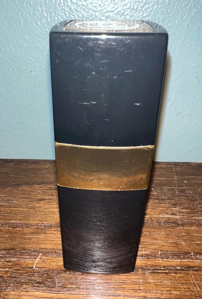 May include: A tall, rectangular black bottle with a smooth, matte finish and a prominent horizontal gold band across the center. The top of the bottle features a circular silver cap with an embossed logo design, and the base has a textured surface.