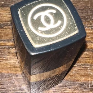 May include: A black lipstick tube with a square profile and a gold-colored band near the base. The top cap features a white interlocking double C logo centered within a square gold-toned inlay. The exterior surface shows visible fine surface scratches.