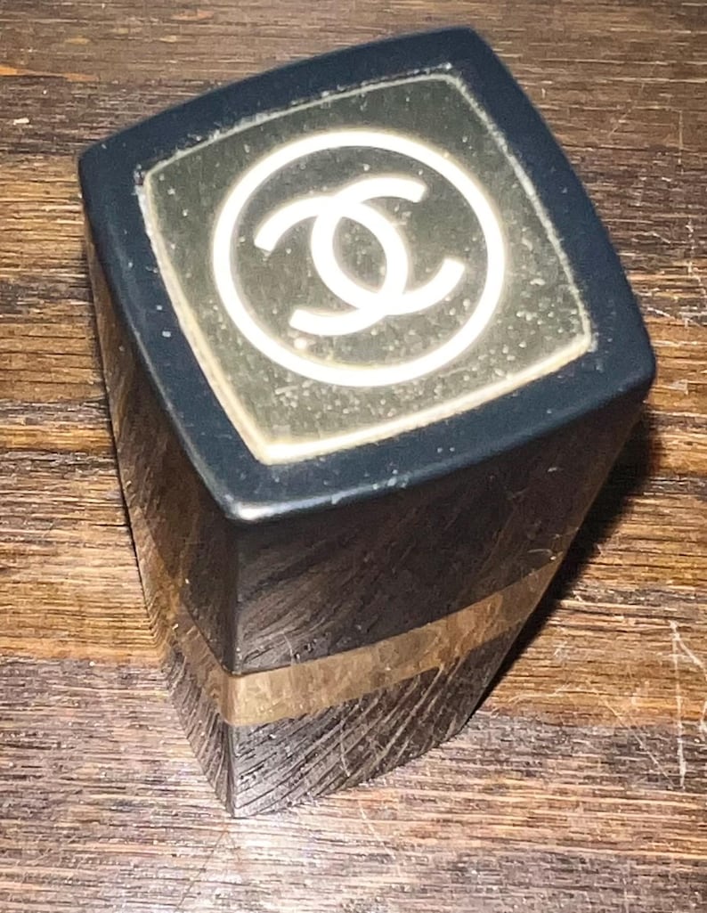 May include: A black lipstick tube with a square profile and a gold-colored band near the base. The top cap features a white interlocking double C logo centered within a square gold-toned inlay. The exterior surface shows visible fine surface scratches.