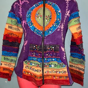 May include: A purple zip-up hoodie with a colorful embroidered design on the front. The sleeves are made of different colored fabric strips and the hoodie has a rainbow pattern on the bottom.