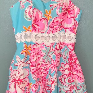 May include: A sleeveless dress featuring a vibrant floral pattern in shades of bright pink, light blue, and orange on a light blue background. The dress includes a white lace-like trim detail around the waist and thin spaghetti straps.