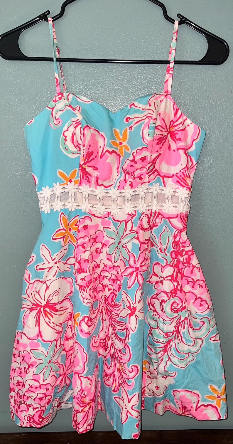 May include: A sleeveless dress featuring a vibrant floral pattern in shades of bright pink, light blue, and orange on a light blue background. The dress includes a white lace-like trim detail around the waist and thin spaghetti straps.