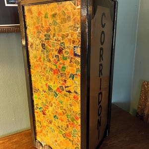 May include: A rectangular, vintage-style display case with a black frame. The front panel is filled with a collage of colorful postage stamps. The word "CORRIDONI" is printed vertically on the side. The case is displayed on a wooden surface.