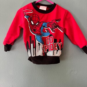 K&ouml;nnte beinhalten: Ein rotes Langarm-Sweatshirt mit einer Grafik von Spider-Man in dynamischer Pose &uuml;ber einer Stadtsilhouette. Der Text "GO SPIDEY" ist in wei&szlig;en Buchstaben aufgedruckt. Das Kleidungsst&uuml;ck hat einen schwarzen gerippten Rundhalsausschnitt, schwarze B&uuml;ndchen und einen schwarzen Saum.