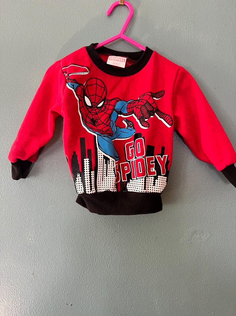 K&ouml;nnte beinhalten: Ein rotes Langarm-Sweatshirt mit einer Grafik von Spider-Man in dynamischer Pose &uuml;ber einer Stadtsilhouette. Der Text "GO SPIDEY" ist in wei&szlig;en Buchstaben aufgedruckt. Das Kleidungsst&uuml;ck hat einen schwarzen gerippten Rundhalsausschnitt, schwarze B&uuml;ndchen und einen schwarzen Saum.