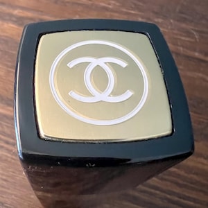 May include: A square-topped black lipstick case featuring a gold-toned metal cap. The top of the cap displays a white circular outline surrounding a white interlocking double C logo, centered on the brushed gold surface of the square lid.
