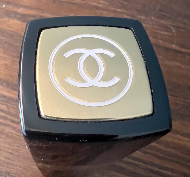 May include: A square-topped black lipstick case featuring a gold-toned metal cap. The top of the cap displays a white circular outline surrounding a white interlocking double C logo, centered on the brushed gold surface of the square lid.