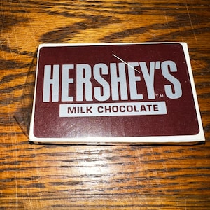 May include: A brown rectangular box with the text "HERSHEY'S" in white letters on a silver background. The text "MILK CHOCOLATE" is also in white letters on a silver background. The text "T.M." is in white letters on a silver background.