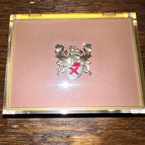 May include: A gold-framed, pink-colored box with a gold crest featuring a red cross inside. The crest is surrounded by gold leaves and a crown.