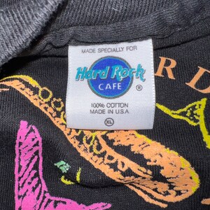 May include: Black t-shirt featuring a saxophone graphic in vibrant colours and the Hard Rock Cafe logo. The label states "Made Specially For" and "100% Cotton Made in U.S.A." The shirt is size XL.