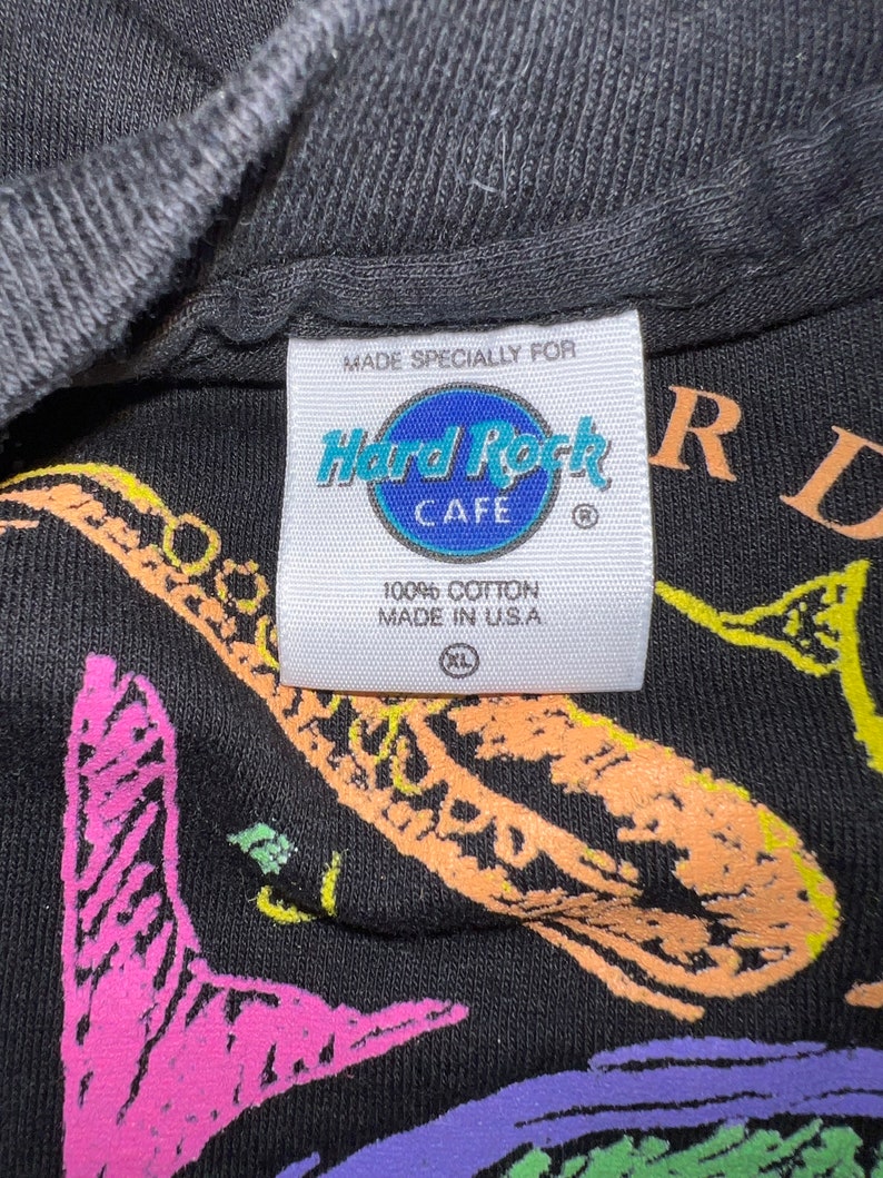 May include: Black t-shirt featuring a saxophone graphic in vibrant colours and the Hard Rock Cafe logo. The label states "Made Specially For" and "100% Cotton Made in U.S.A." The shirt is size XL.