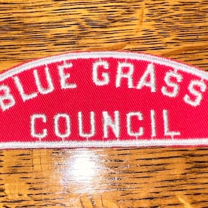May include: A red embroidered patch with the words "BLUE GRASS COUNCIL" in white, arched across the top and bottom. The patch has a white border and is set against a wooden background.