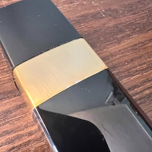 May include: A rectangular object featuring a matte black top section, a central metallic gold band, and a glossy black lower portion. The design is sleek and geometric, with a clear transition between the three distinct color and texture segments.