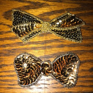 May include: Two gold sequined bows. The bows are made of small, round sequins sewn together in a bow shape. The bows are on a wooden surface.