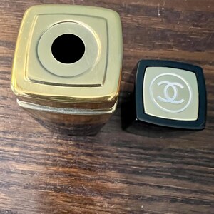 May include: A square-shaped lipstick tube with a metallic gold top and a black base. The detached black cap features a gold square inlay with a white interlocking double C logo centered on the top surface. The lipstick tube is open at the top.