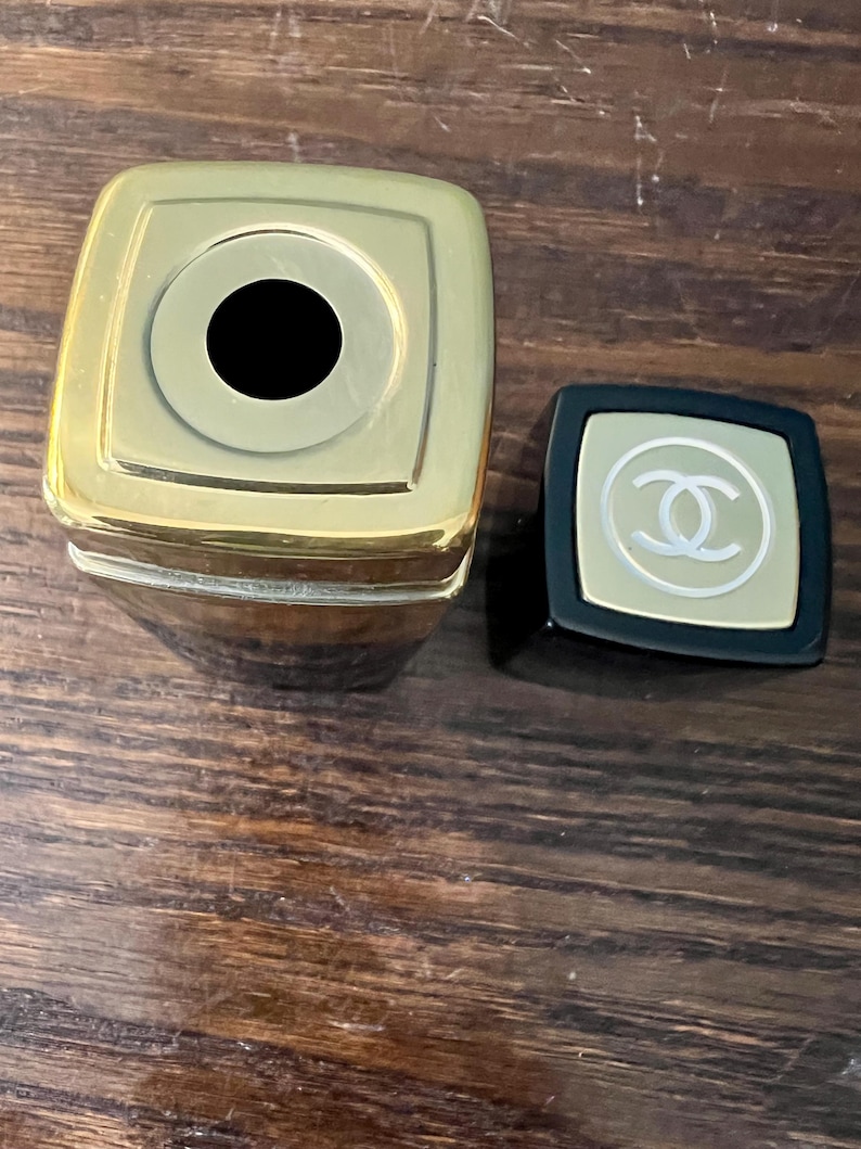 May include: A square-shaped lipstick tube with a metallic gold top and a black base. The detached black cap features a gold square inlay with a white interlocking double C logo centered on the top surface. The lipstick tube is open at the top.
