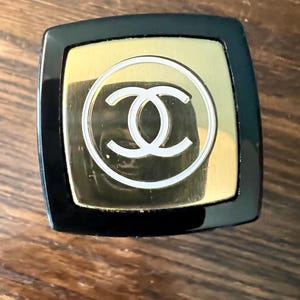 May include: A square-shaped cosmetic cap featuring a glossy black outer border and a central gold-toned square plate. The gold plate displays a white circular outline surrounding a white interlocking double C logo centered on the top surface.