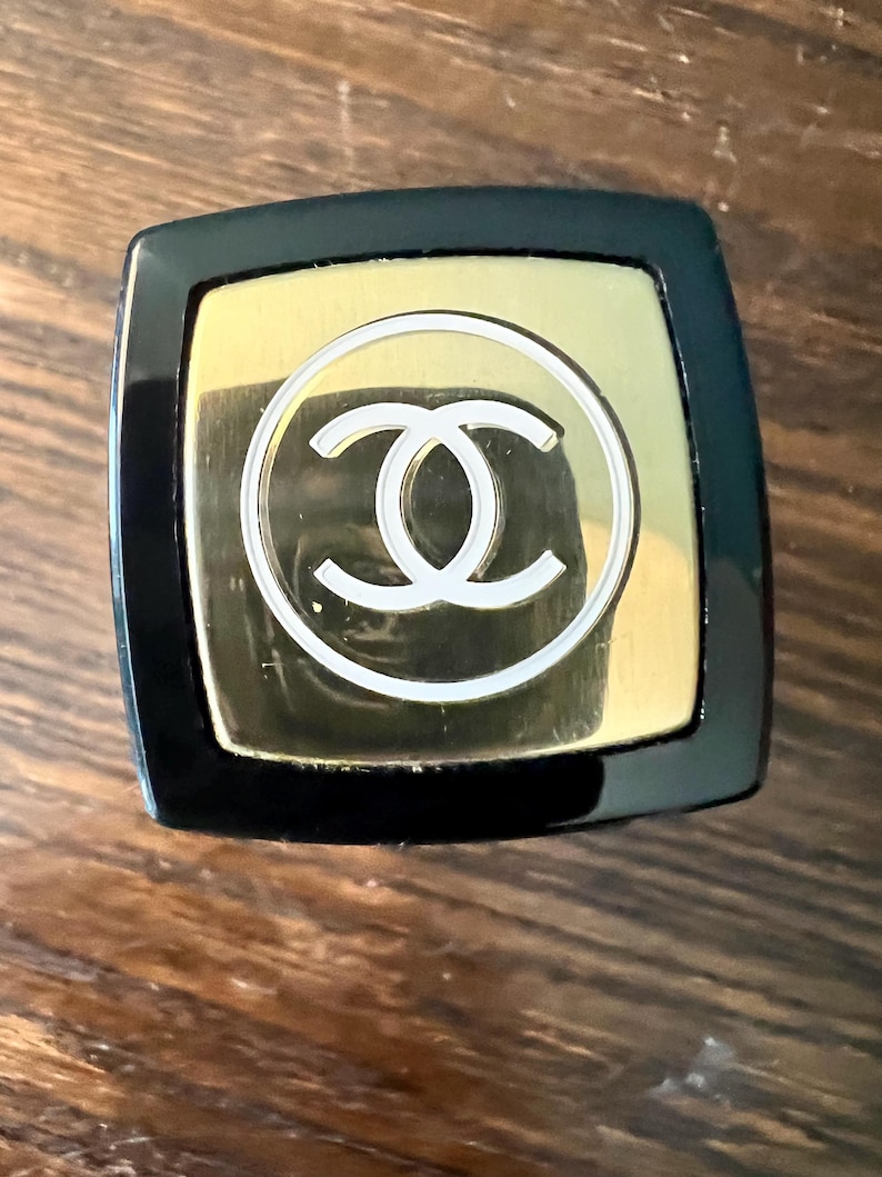 May include: A square-shaped cosmetic cap featuring a glossy black outer border and a central gold-toned square plate. The gold plate displays a white circular outline surrounding a white interlocking double C logo centered on the top surface.