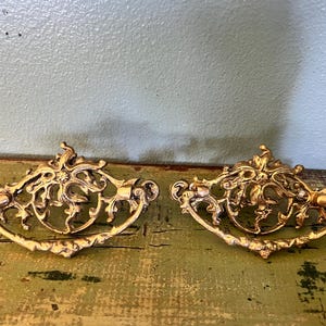 May include: Two ornate, gold-colored drawer pulls with intricate scrollwork and a curved handle. The hardware is likely vintage or antique, adding a touch of elegance to furniture. The pulls are displayed on a distressed, green-painted wooden surface.
