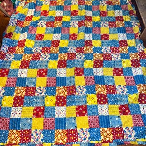 May include: A colorful patchwork quilt with a grid pattern. The quilt features squares of red, yellow, and blue fabrics with various floral and geometric patterns. The quilt is spread out on a wooden surface.