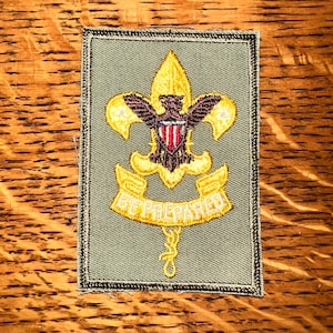 May include: A rectangular patch with a green background and a gold border. The patch features a gold fleur-de-lis with a brown eagle and a red and white shield. The words "BE PREPARED" are written on a gold banner.