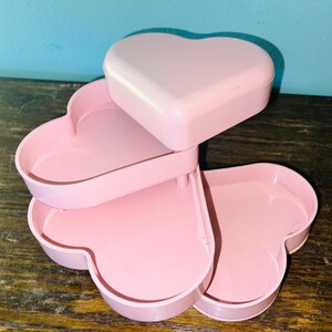 May include: A light pink, multi-tiered storage organizer featuring three heart-shaped trays that rotate outward. The top tray includes a solid heart-shaped lid, while the lower two trays provide open storage compartments for small items.