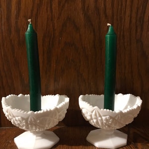 May include: Two white milk glass candle holders with a scalloped edge and a pedestal base. Each holder has a dark green candle.