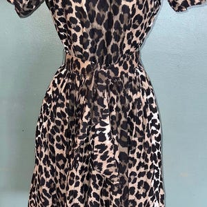 May include: A short-sleeved dress featuring a tan and black leopard print pattern. The garment includes a fitted waist with a matching fabric tie belt at the back and a flared skirt that falls to the knees, displayed on a mannequin form.