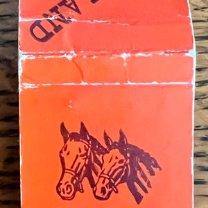 May include: An orange matchbox with the word KEENELAND in bold, dark red letters. The matchbox features two horse heads in a dark red color. The text "Please Close Cover Before Striking" is visible.
