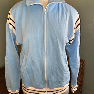 May include: Light blue zip-up jacket with white and navy stripes on the sleeves and hem. The jacket has a high collar, a white zipper, and two front pockets. The jacket is displayed on a mannequin.
