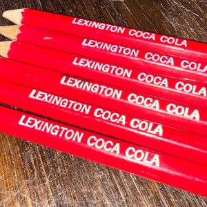 May include: A set of six red wooden pencils, each featuring the white printed text LEXINGTON COCA COLA. The pencils have sharpened graphite tips and a hexagonal barrel shape, arranged in a parallel stack to display the branding clearly.