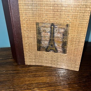 May include: A square photo album with a textured tan woven cover and a dark brown spine. The center features a square inset graphic depicting the Eiffel Tower with the word Paris written in script, framed by decorative black metal-like scrollwork.