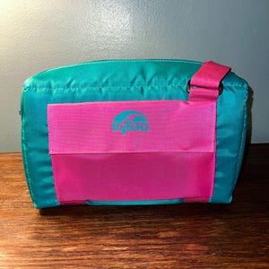 May include: A teal and pink Igloo cooler bag with a shoulder strap. The bag has a front pocket with the Igloo logo.