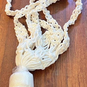 May include: A cream-colored macrame plant hanger, with a woven net design and a bundled base. The hanger is made of thick, soft rope and is designed to hold a plant pot. The hanger is approximately 40 inches long.