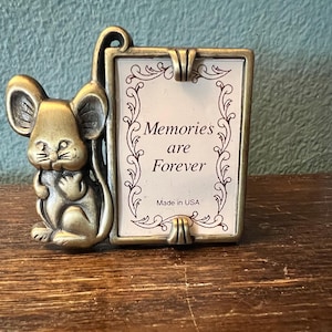 May include: A brass-toned metal picture frame featuring a sculpted mouse figure on the left side. The rectangular frame holds a white insert with decorative scrollwork borders and the printed text Memories are Forever and Made in USA.