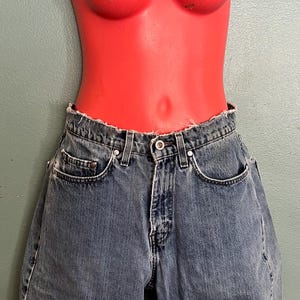 May include: Blue denim shorts with a frayed hem are displayed on a red mannequin torso. The shorts have a button closure, pockets, and belt loops. The mannequin has a smooth, glossy finish.