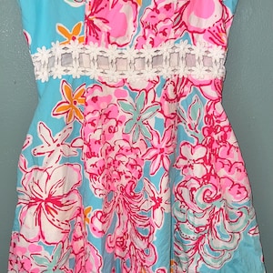 May include: A sleeveless dress featuring a vibrant floral pattern in shades of bright pink, light blue, white, and orange. The garment includes a horizontal white lace trim detail at the waist and thin shoulder straps with a sweetheart neckline.