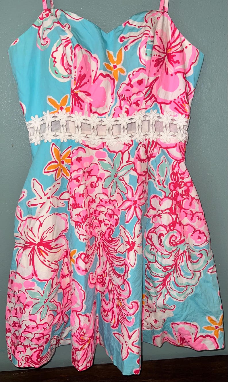 May include: A sleeveless dress featuring a vibrant floral pattern in shades of bright pink, light blue, white, and orange. The garment includes a horizontal white lace trim detail at the waist and thin shoulder straps with a sweetheart neckline.
