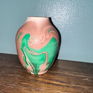 May include: A small, rounded vase featuring a marbled pattern of muted pink and vibrant green swirls. The vessel has a smooth, matte-like finish with a narrow opening at the top and a wider, bulbous body that tapers toward the base.