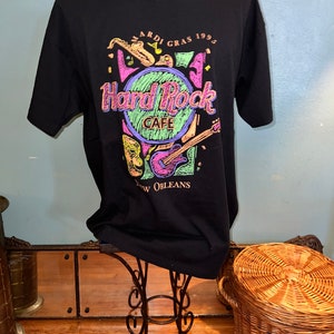 May include: Black t-shirt with a Hard Rock Cafe design, featuring the text "Mardi Gras 1995 New Orleans." The graphic includes a saxophone, guitar, and mask in vibrant colours. The shirt is displayed on a mannequin.