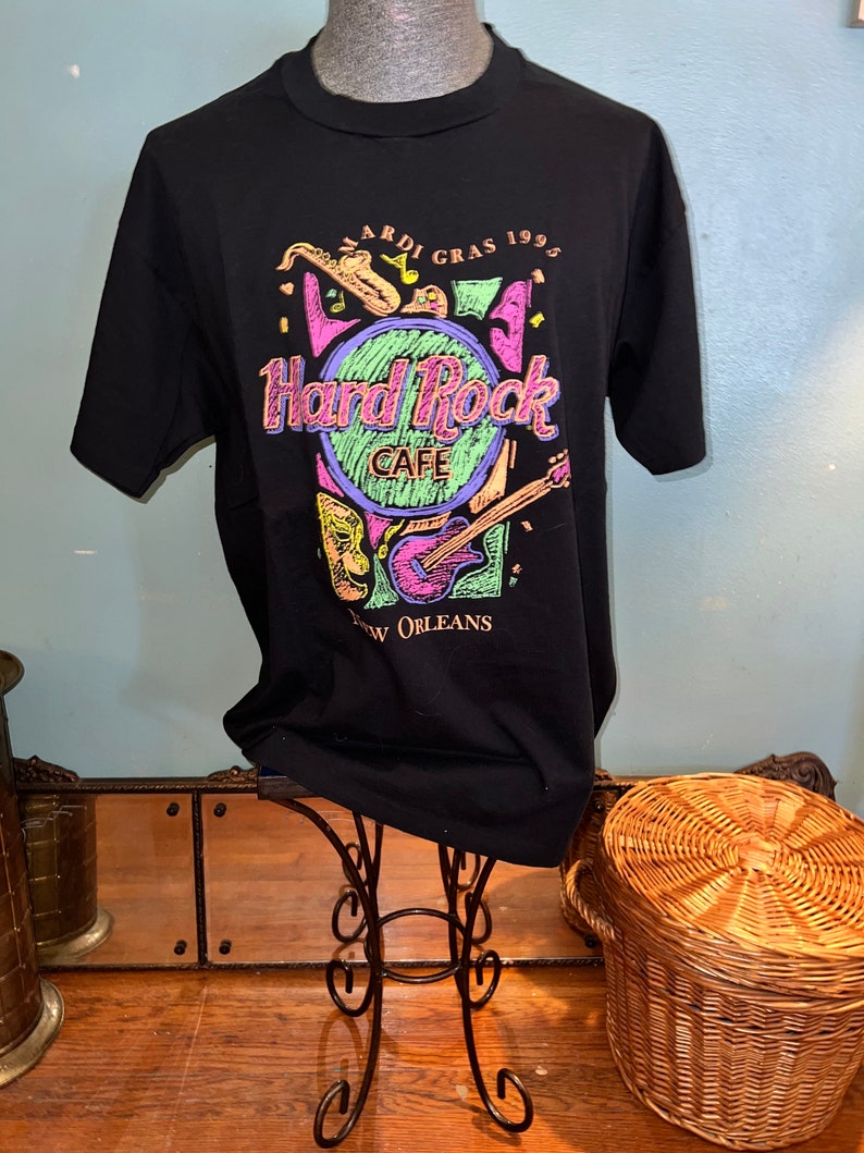 May include: Black t-shirt with a Hard Rock Cafe design, featuring the text "Mardi Gras 1995 New Orleans." The graphic includes a saxophone, guitar, and mask in vibrant colours. The shirt is displayed on a mannequin.