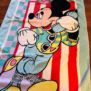 May include: A colorful beach towel featuring Mickey Mouse and Minnie Mouse. The towel has a light blue border and a central design with red and white stripes. Mickey Mouse is depicted in a vibrant outfit with sunglasses. The words "Mickey & Minnie" are also visible.