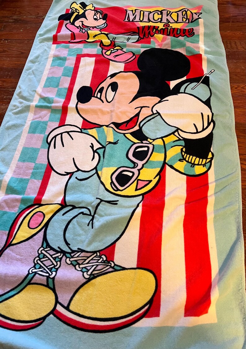 May include: A colorful beach towel featuring Mickey Mouse and Minnie Mouse. The towel has a light blue border and a central design with red and white stripes. Mickey Mouse is depicted in a vibrant outfit with sunglasses. The words "Mickey & Minnie" are also visible.