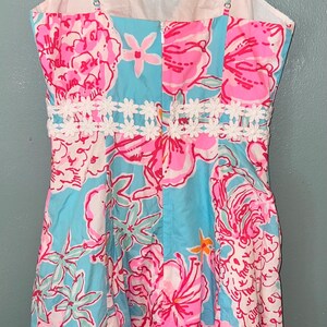 May include: A sleeveless dress featuring a vibrant floral pattern in shades of bright pink, white, and light blue. The dress has a sweetheart neckline, thin spaghetti straps, and a white lace trim detail around the waist with a center back zipper.