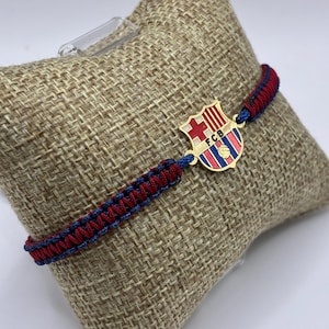 May include: A red and blue braided bracelet with a gold-colored FC Barcelona emblem. The emblem features the team's crest with a cross, stripes, and a soccer ball. The bracelet is displayed on a tan, textured surface.