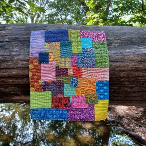 May include: A colorful, square patchwork quilt with a variety of patterned fabrics in shades of blue, green, pink, purple, and orange. The quilt is hand-stitched with visible thread and is displayed on a weathered log.
