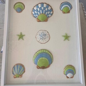 以下が含まれることがあります： 絵画の額縁には、塗装された貝殻とヒトデが描かれています。貝殻は青、緑、白の色合いで、金色のアクセントが付いています。配置には、サンドダラーと2つの緑色のヒトデが含まれています。