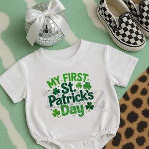 Può includere: Body da bambino bianco con testo verde "MY FIRST St. Patrick's Day" e grafica a trifogli. Una palla da discoteca argentata con un fiocco bianco e scarpe slip-on a scacchi bianchi e neri sono sullo sfondo.