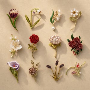 May include: A collection of twelve floral brooches featuring various designs including a purple and green flower, a white bell-shaped bloom, a pearl-accented spray, a daisy, a white blossom, a red rose, a hydrangea, a dark red cluster, a calla lily, a rose, wheat-like stalks, and a pink floral sprig.